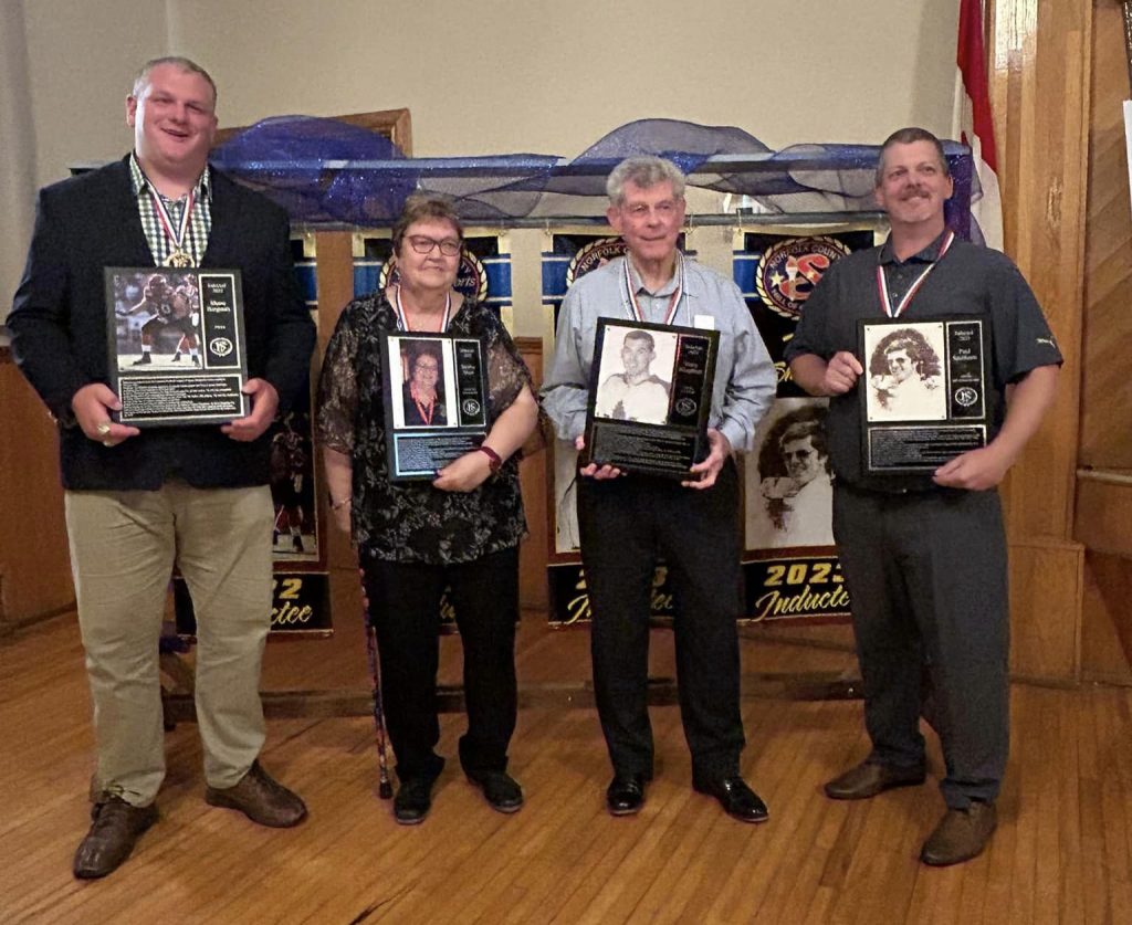 A photo of the inductees from the 2024 Induction Dinner, holding their plaques and standing in front of the banners commemorating their achievements.
